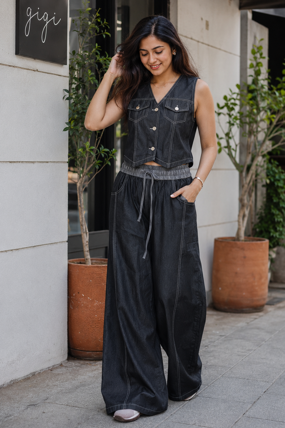 Woman in a black denim vest and wide-leg pants standing outside a building with 'gigi' sign.
